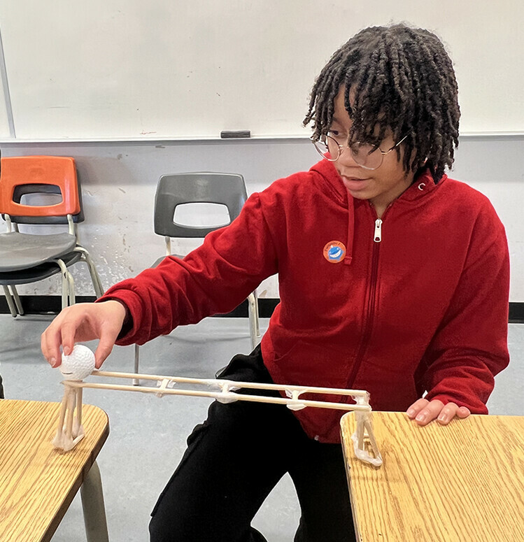 Student rolling a golf ball from desk to desk, using a ramp built themselves.