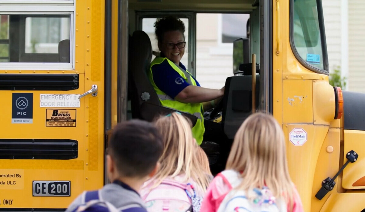 Smiling bus driver sitting in the bus with the door open to children walking in.