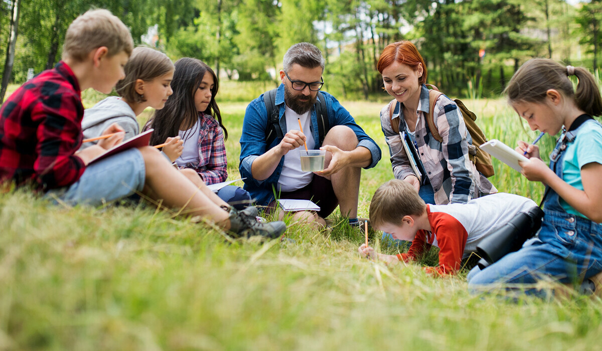 Two adults and six children sit in a grassy field, observing nature and taking notes during an outdoor learning activity.