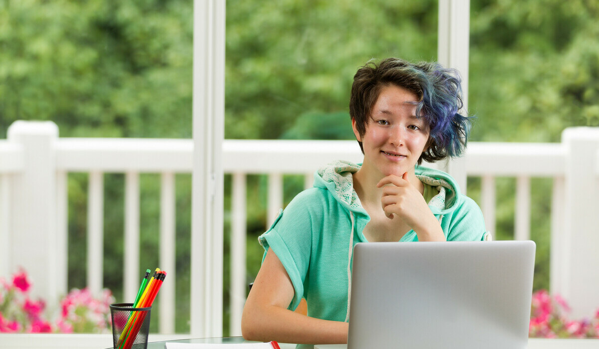 A teen with short, dark hair dyed blue on one side sits at a table with a laptop, notebooks and pencils, smiling in front of a bright window overlooking a green backyard.