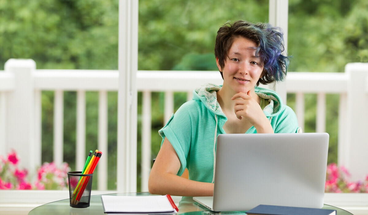 A teen with short, dark hair dyed blue on one side sits at a table with a laptop, notebooks and pencils, smiling in front of a bright window overlooking a green backyard.