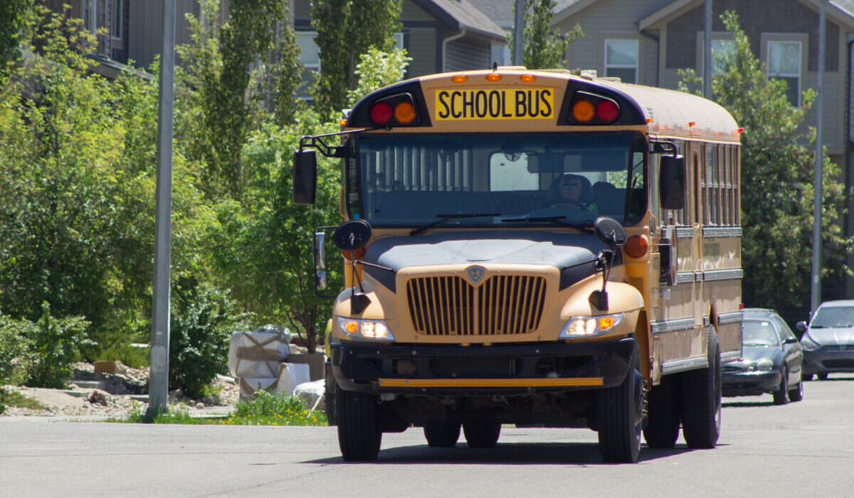 A smiling school bus driver wearing a safety vest sits behind the wheel of a yellow school bus.
