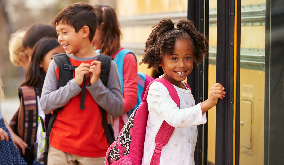 Group of diverse young children boarding the school bus. The girl in front is wearing a pink backpack and white shirt. She is smiling at the camera.
