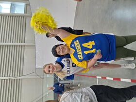 Two students wearing Bert Church Chargers sports jerseys smile while participating in the sports jamboree. One student is holding a yellow pom pom in the air.